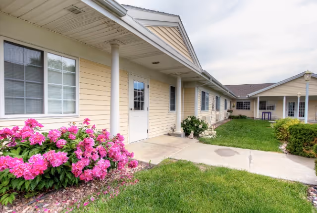 Exterior view of a single-story senior living facility building with beige siding, white trim, and a covered walkway supported by white columns. There are windows along the building and a white door. The foreground features a concrete pathway, green grass, and blooming pink flowers near the building.
