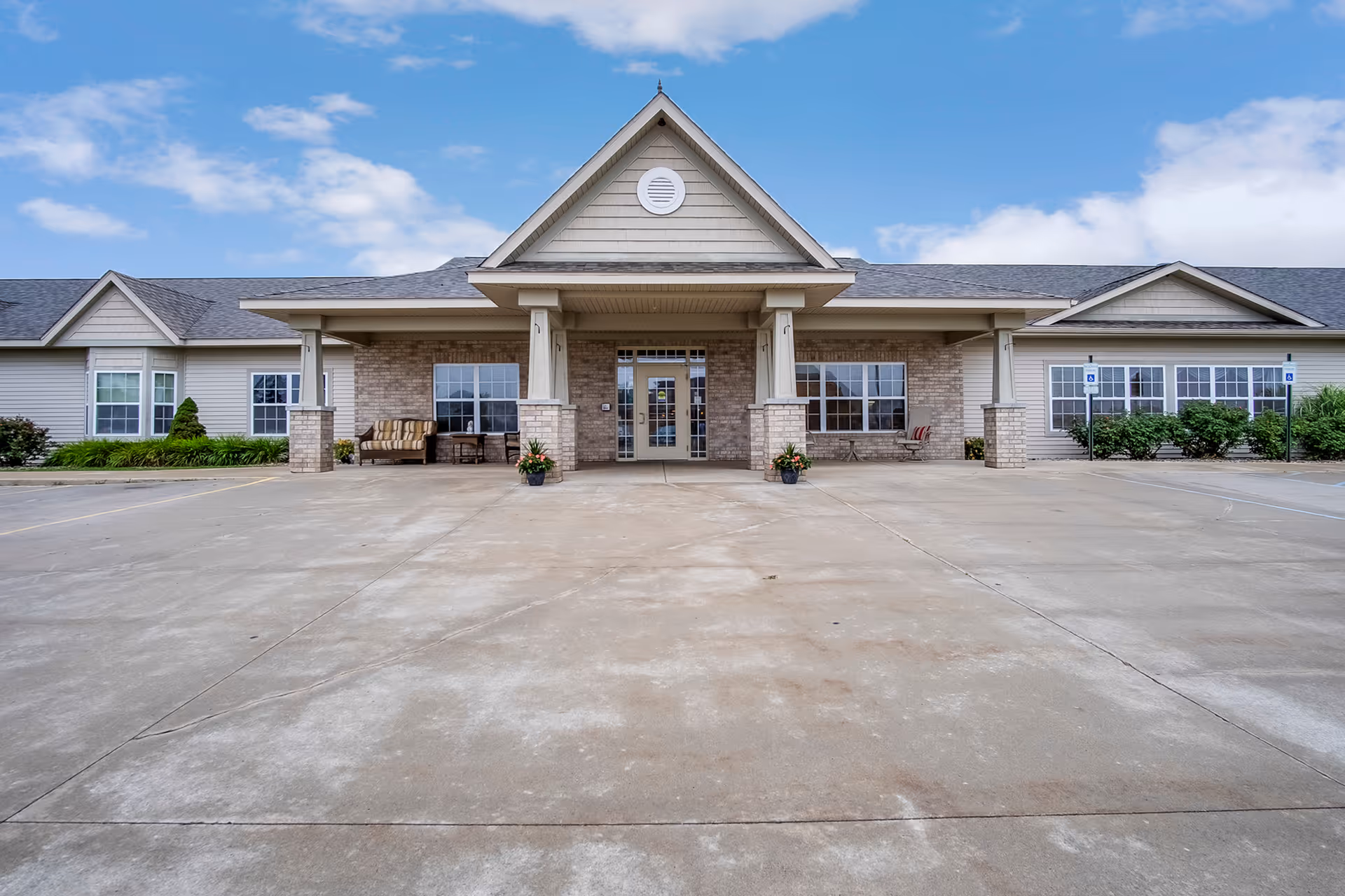 Front exterior and covered entrance of a single-story senior living facility with a wide paved driveway, seating, and potted plants.