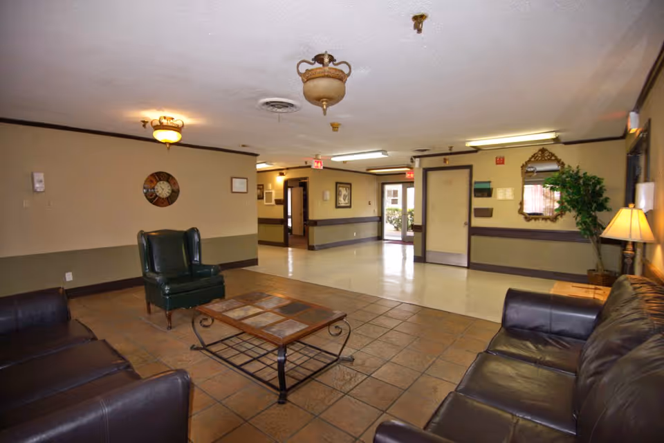 A common area in a nursing and rehabilitation center featuring a tiled floor section with a coffee table and leather sofas, a green armchair, beige walls with dark trim, a wall clock, framed pictures, a mirror, a potted plant, and ceiling light fixtures. There is a hallway and an exit door visible in the background.