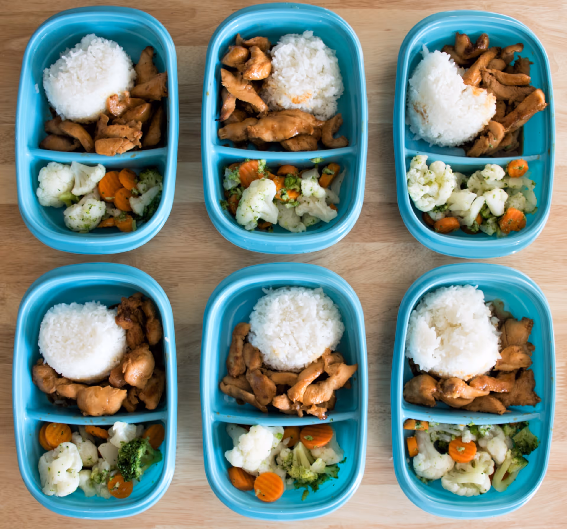 Six turquoise meal trays hold rice, chicken, and vegetables on a wooden table.