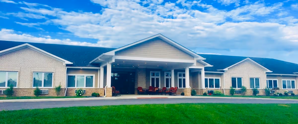 Front exterior view of Autumn Trace Rensselaer Assisted Living building with a covered entrance, several windows, and a well-maintained lawn under a partly cloudy blue sky.