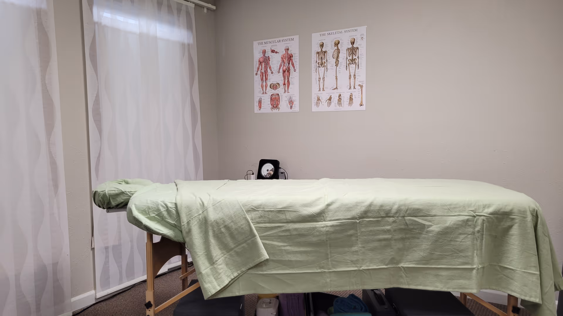 A massage table covered with a green sheet sits in a therapy room with curtains.