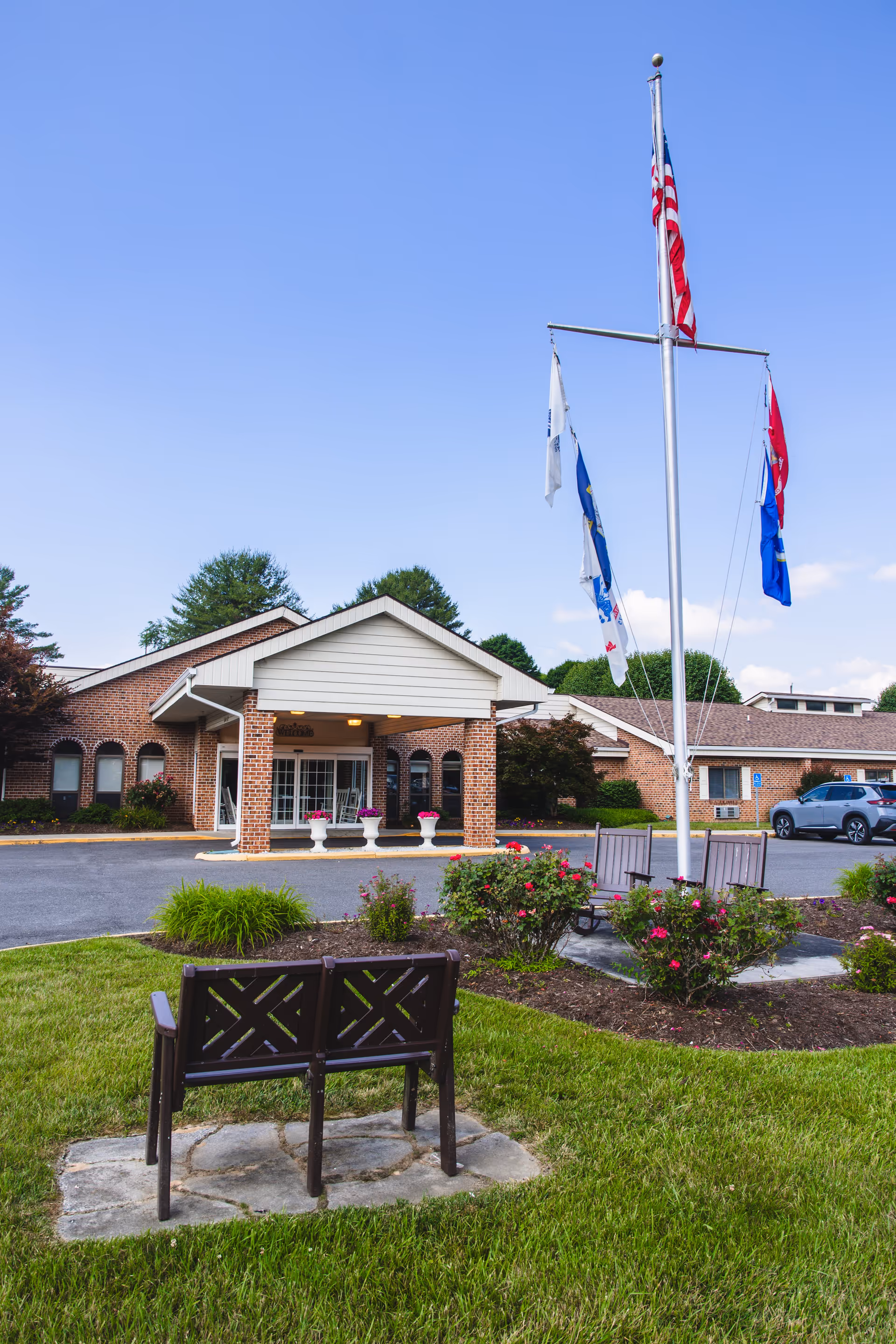 Exterior view of Commonwealth Senior Living At Hillsville showing a brick building with a covered entrance, a flagpole with multiple flags, landscaped garden beds with flowers, and benches on a grassy area under a clear blue sky.