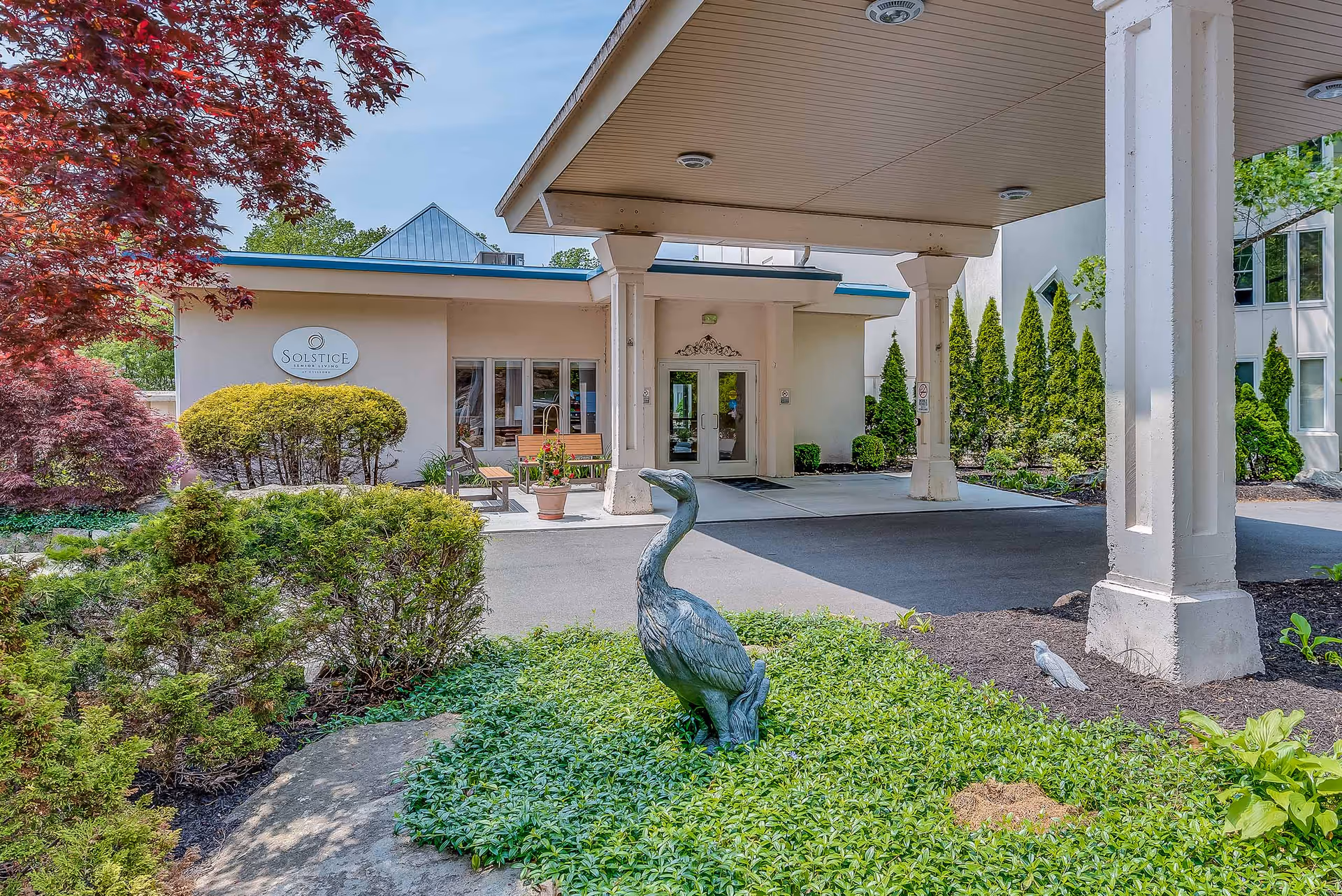 Entrance of a retirement residence with a covered porte-cochere, landscaped beds and a metal heron statue in the foreground.