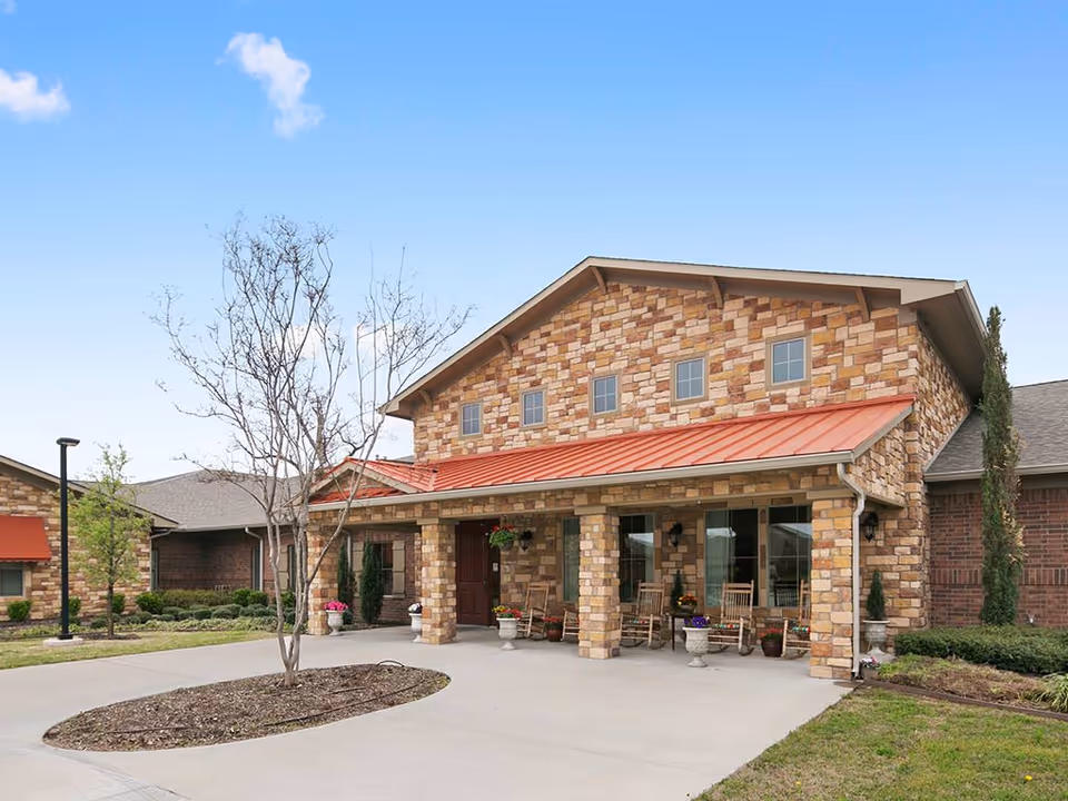 Front entrance of a stone-clad senior living building with a red metal awning, covered porch with rocking chairs and planters, and a circular driveway.