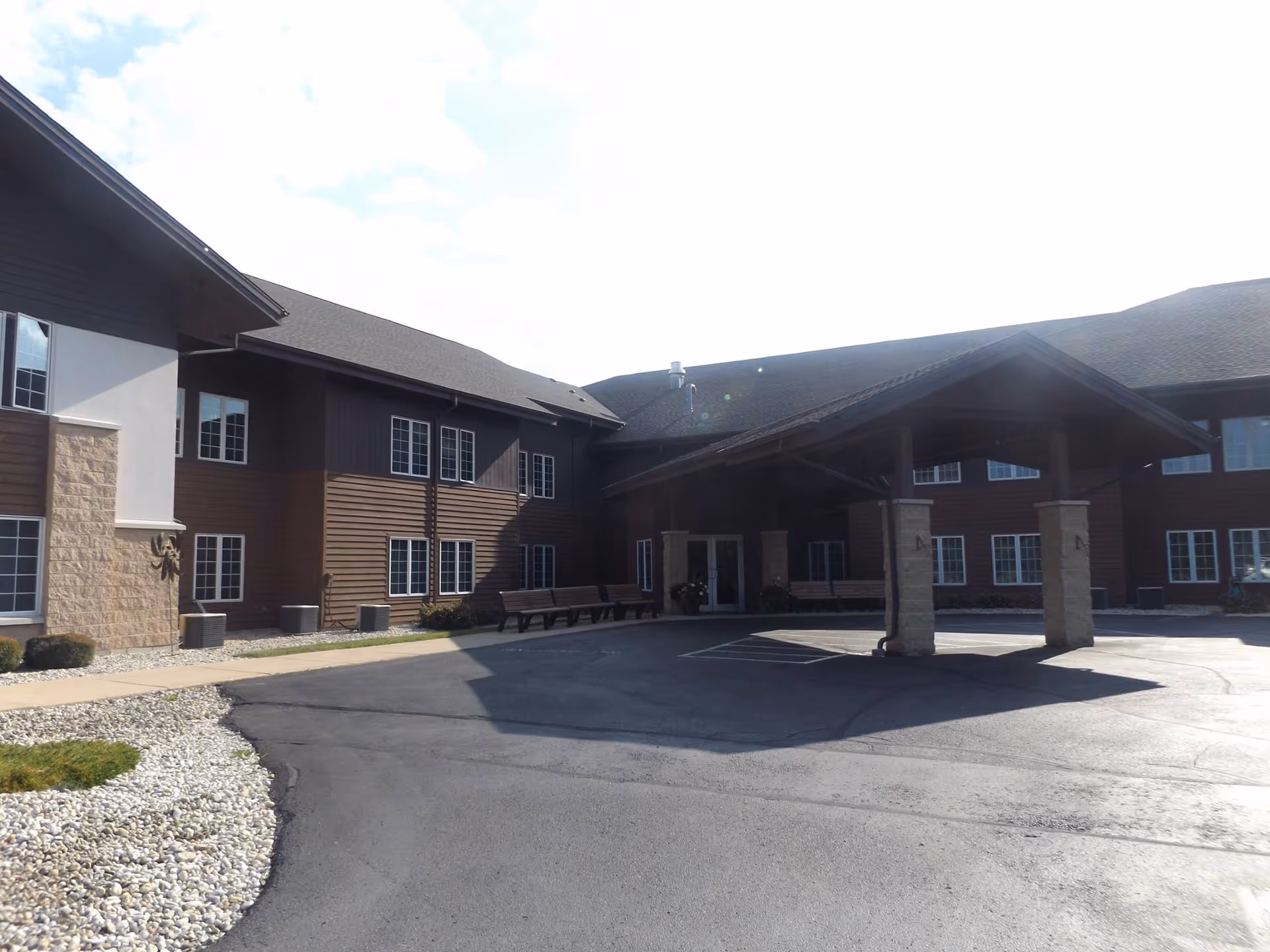 Exterior view of St Clare Friedensheim Assisted Living facility showing a two-story building with brown siding and stone accents. There is a covered entrance supported by stone pillars and benches along the walkway. The area in front is paved with asphalt and landscaped with white rocks and small bushes.
