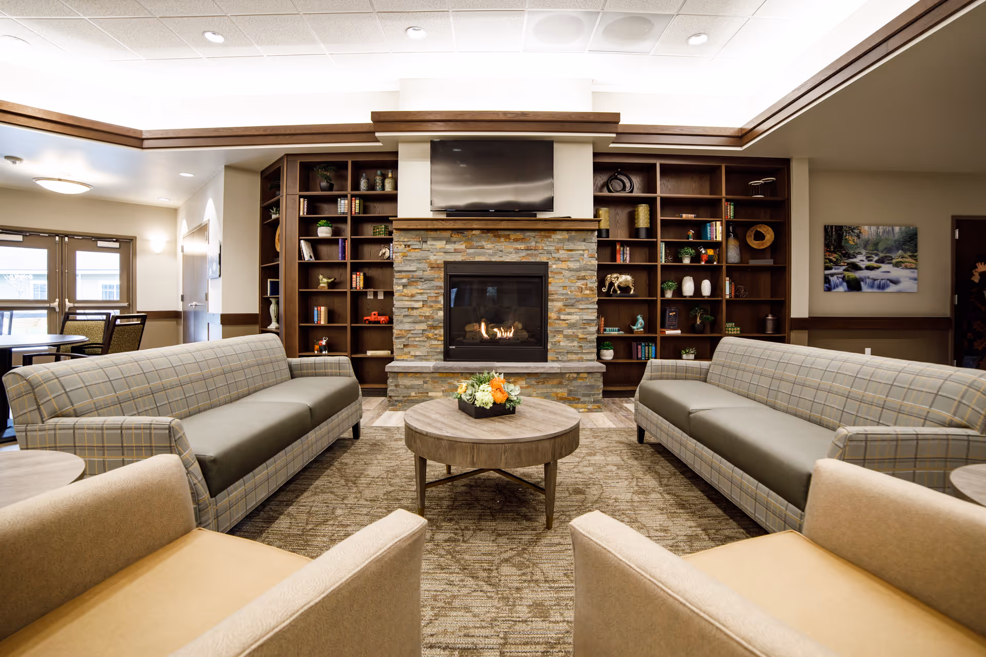 A cozy living room area with two plaid sofas facing each other and two beige armchairs in the foreground. A round wooden coffee table with a flower arrangement sits in the center. In the background, there is a stone fireplace with a flat-screen TV mounted above it, flanked by built-in wooden bookshelves filled with books and decorative items. The room has a warm and inviting atmosphere with soft lighting and a carpeted floor.