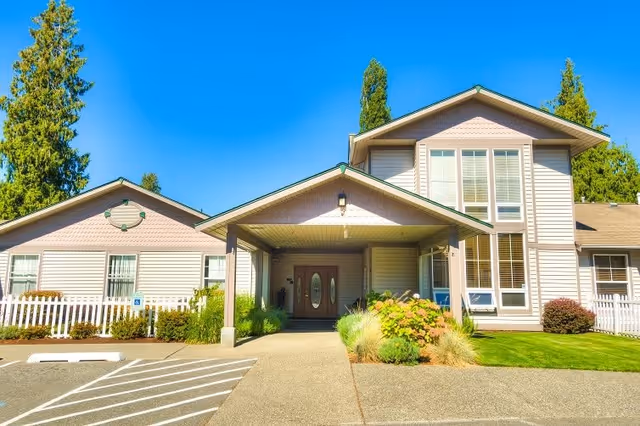 Exterior front view of a senior living facility building with a covered entrance, double doors, large windows, manicured lawn, and surrounding trees under a clear blue sky.