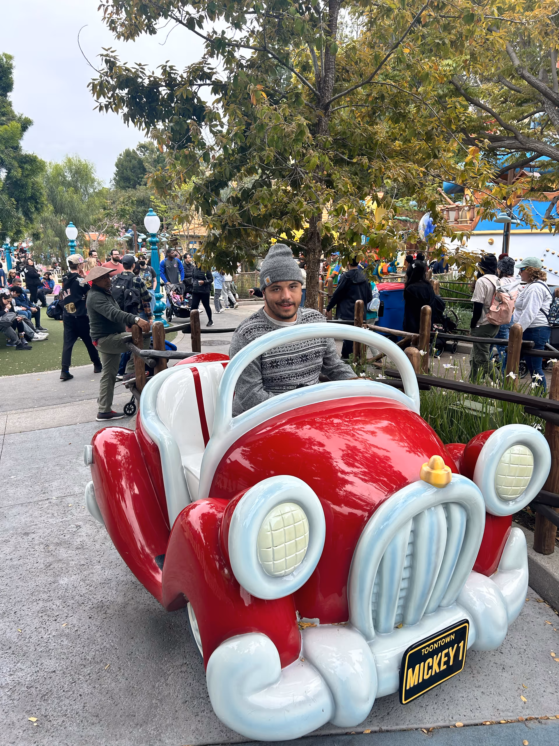 A person sits in a bright red vintage-style children's car ride in a crowded outdoor park.