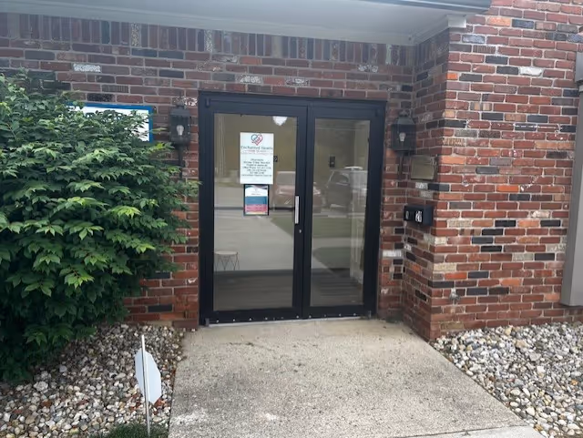 Brick building entrance with glass double doors, concrete walkway, and surrounding rocks.