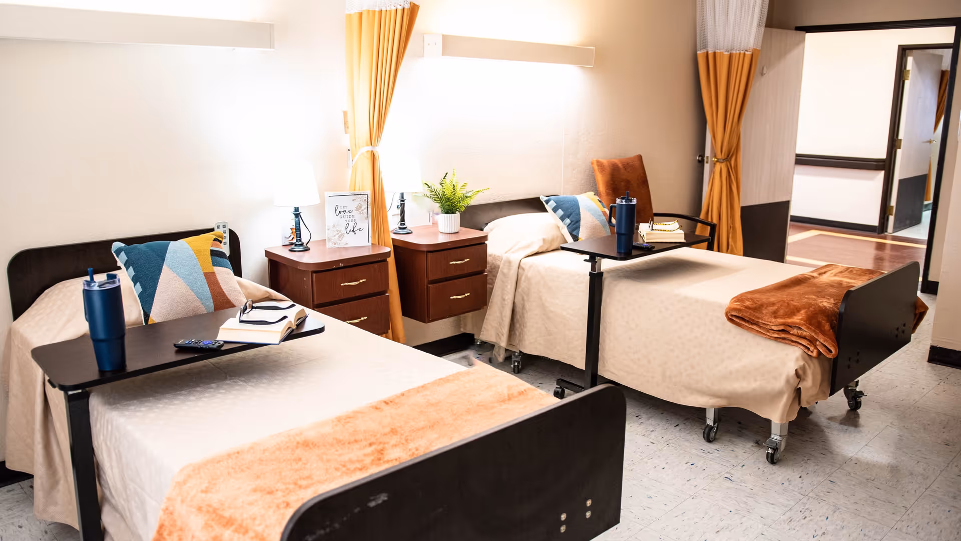 A bright and clean bedroom in a senior living facility with two single beds, each with a beige bedspread, colorful pillows, and a brown throw blanket. Each bed has a black overbed table with a blue tumbler and books. Between the beds are two wooden nightstands with lamps, a small plant, and a framed decorative sign. The room has light-colored walls, yellow curtains, and an open doorway leading to a hallway.