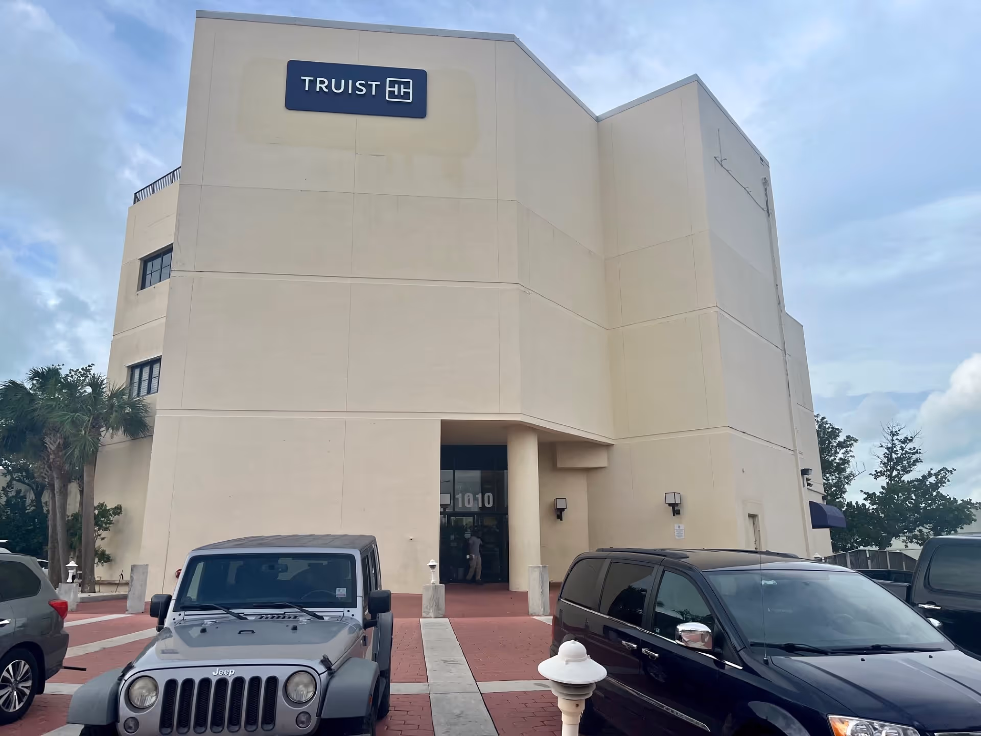 Beige building exterior with a front entrance sign and parked cars in the lot.