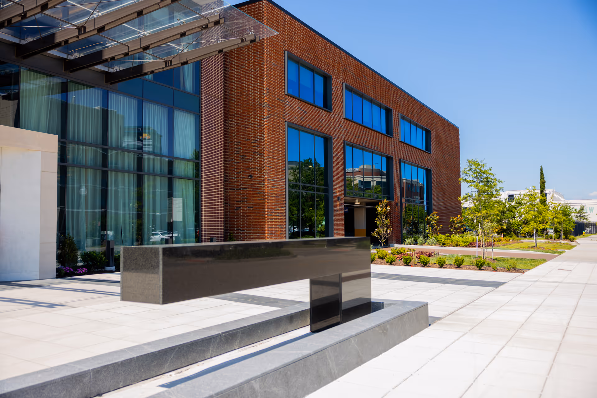 Modern brick building with large glass windows reflecting the blue sky, surrounded by a paved walkway and landscaped greenery under a clear blue sky.