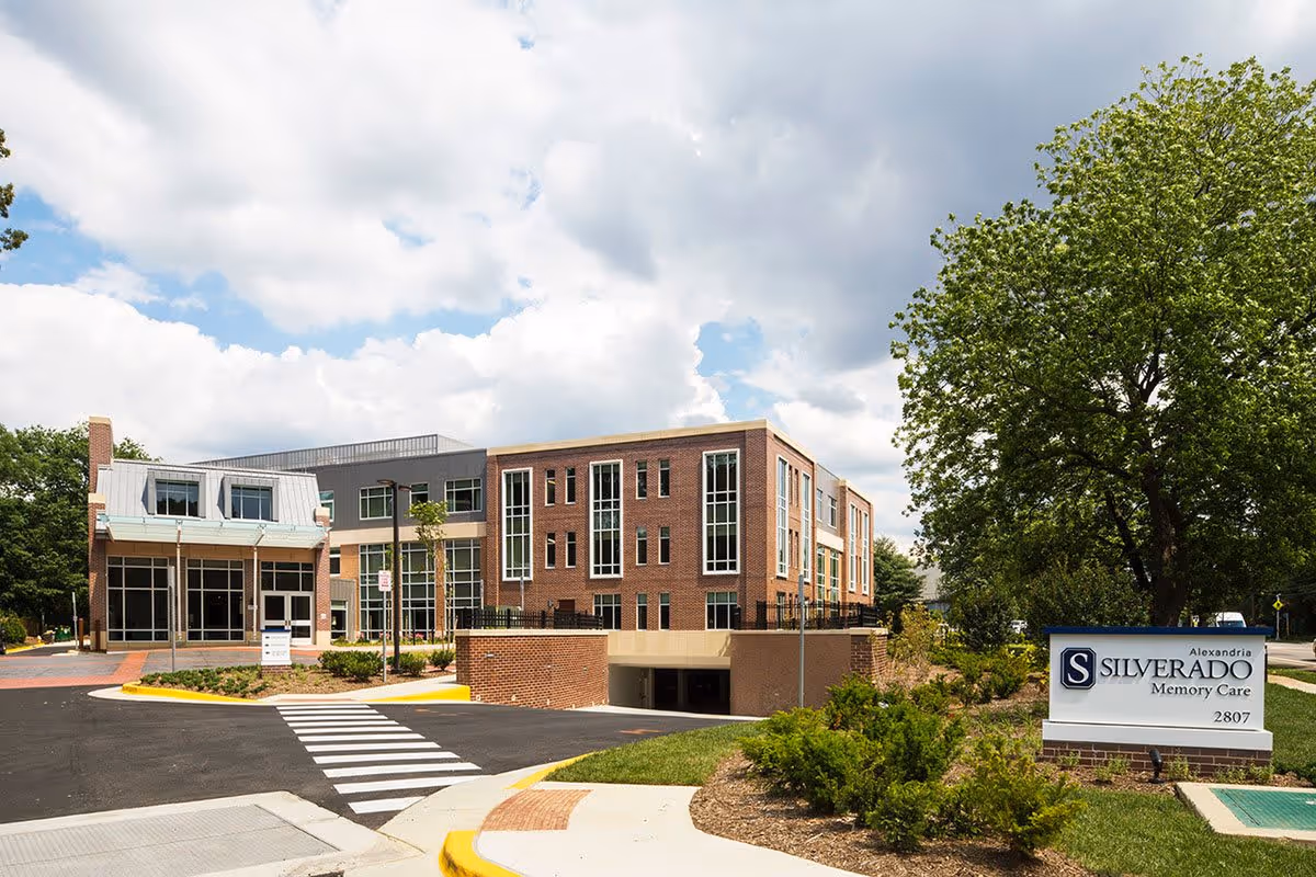 Exterior view of Silverado Alexandria Memory Care Community building with a brick facade, large windows, and a driveway with a pedestrian crosswalk. There is a sign in front displaying the facility name and address, surrounded by landscaping and trees under a partly cloudy sky.