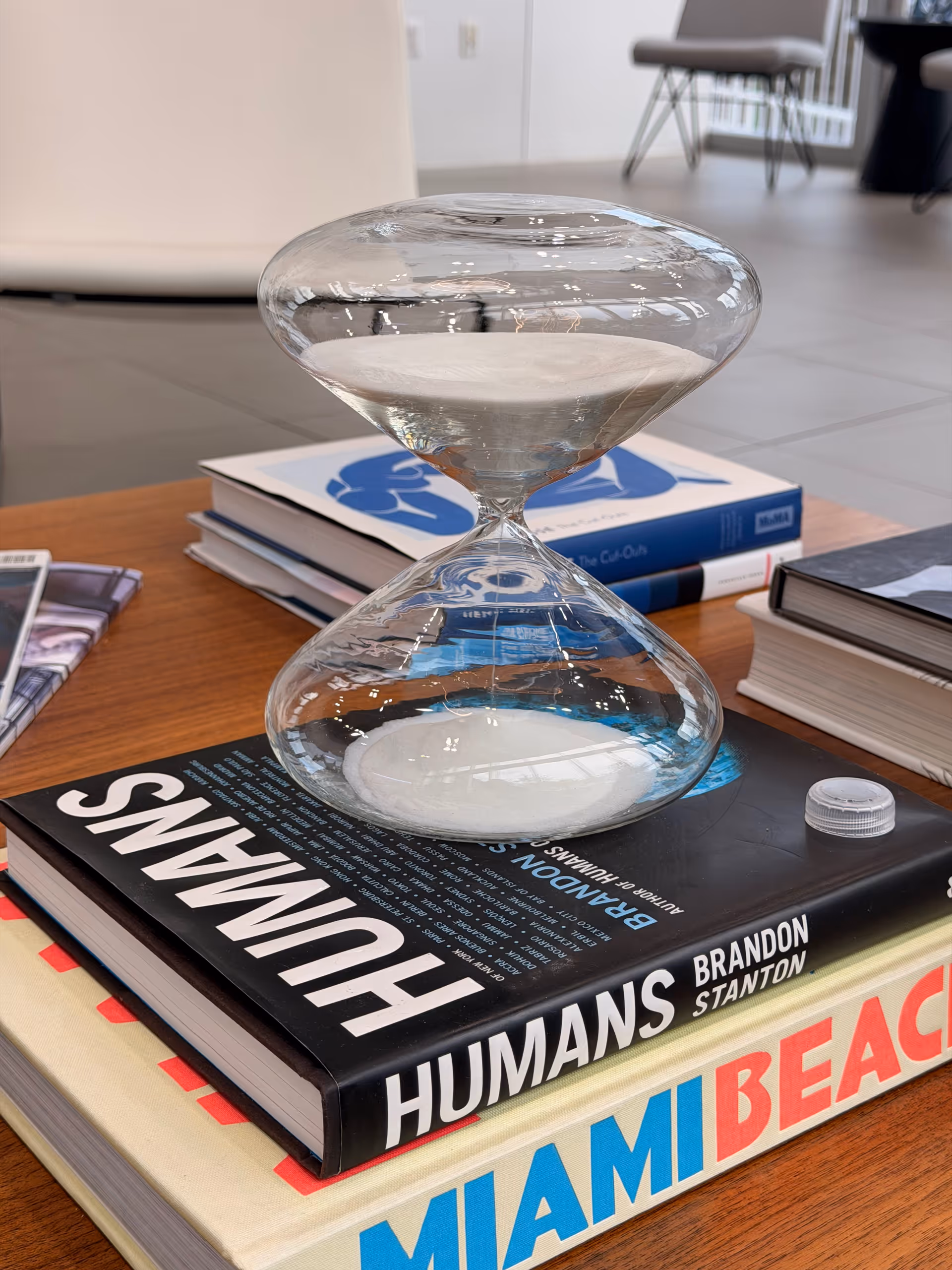 Glass hourglass atop stacked books on a wooden table in a bright living area.