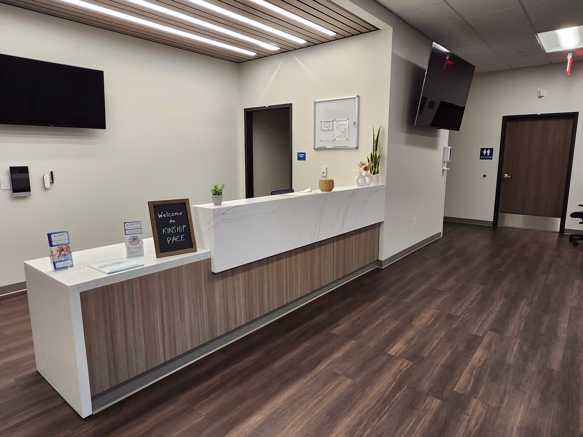 Reception desk in Kinship PACE facility lobby with wood floors and wall-mounted TVs.