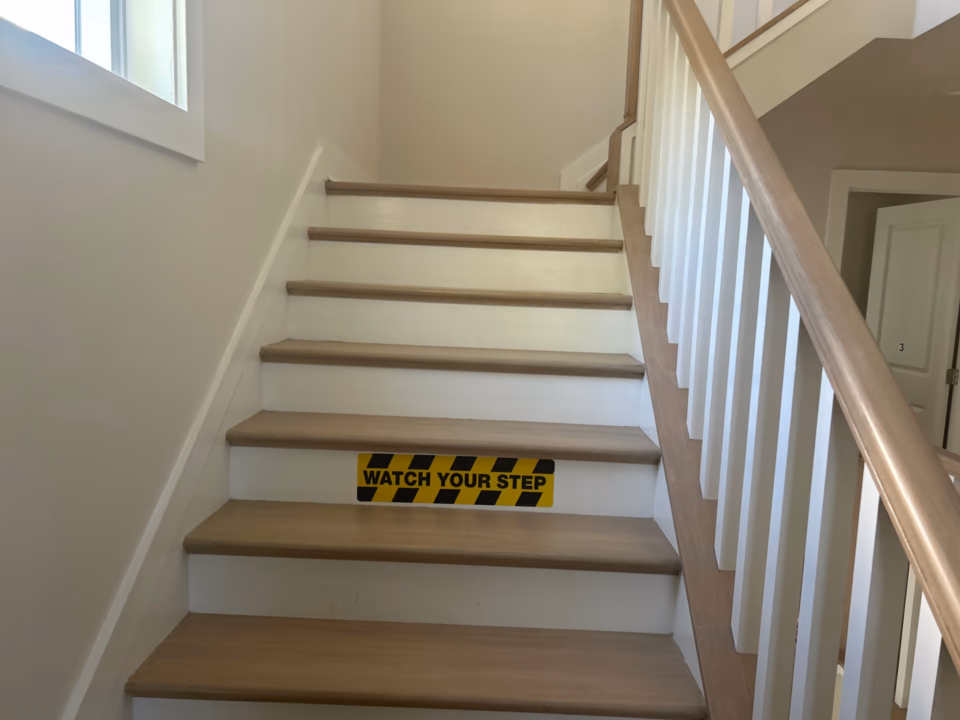 Interior staircase with wooden steps, white balusters, and a yellow 'Watch Your Step' sign.