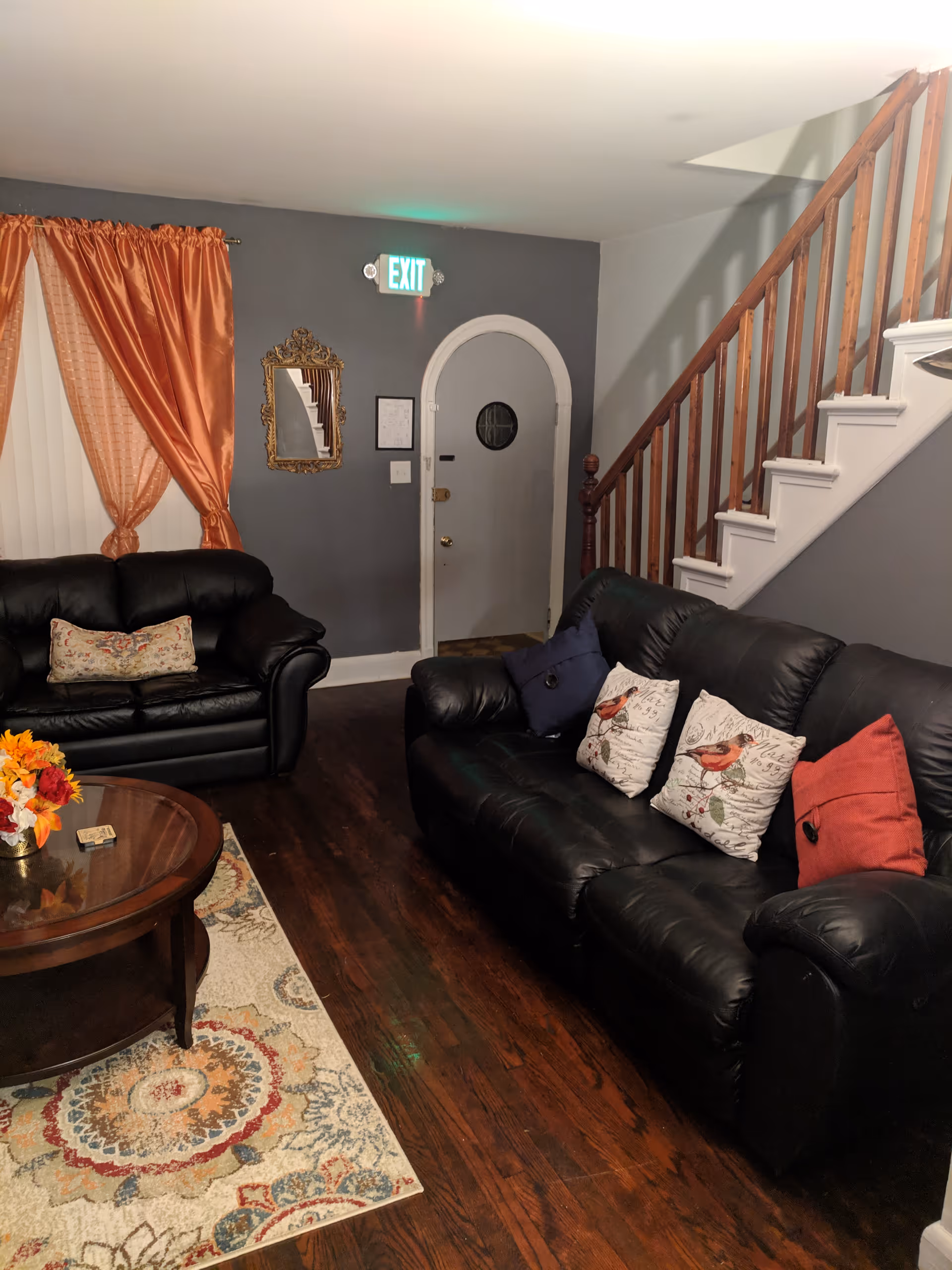 A cozy living room with dark leather sofas adorned with decorative pillows, a wooden coffee table with a flower arrangement, a patterned area rug, orange curtains on the window, a gray wall with a decorative mirror, an exit sign above a white door with a small round window, and a wooden staircase with white risers.