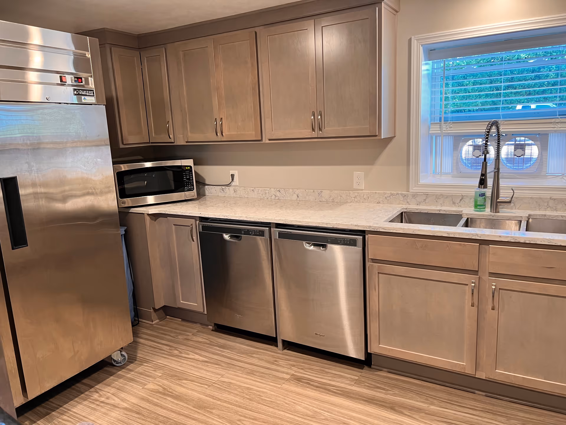 A kitchen area with light wood cabinets, a stainless steel refrigerator, a microwave on the countertop, two stainless steel dishwashers, a double sink with a modern faucet, and a window with blinds above the sink. The floor has a wood-like finish.
