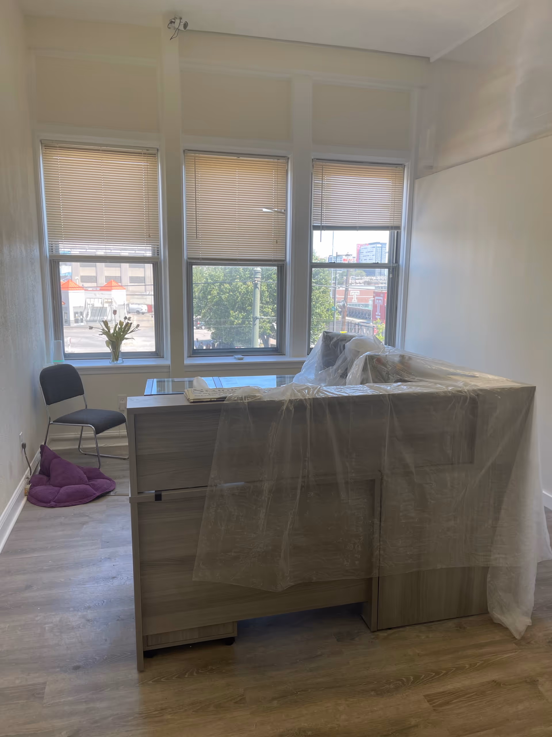 Unfinished interior room with a wooden desk covered in plastic, three windows, and a chair