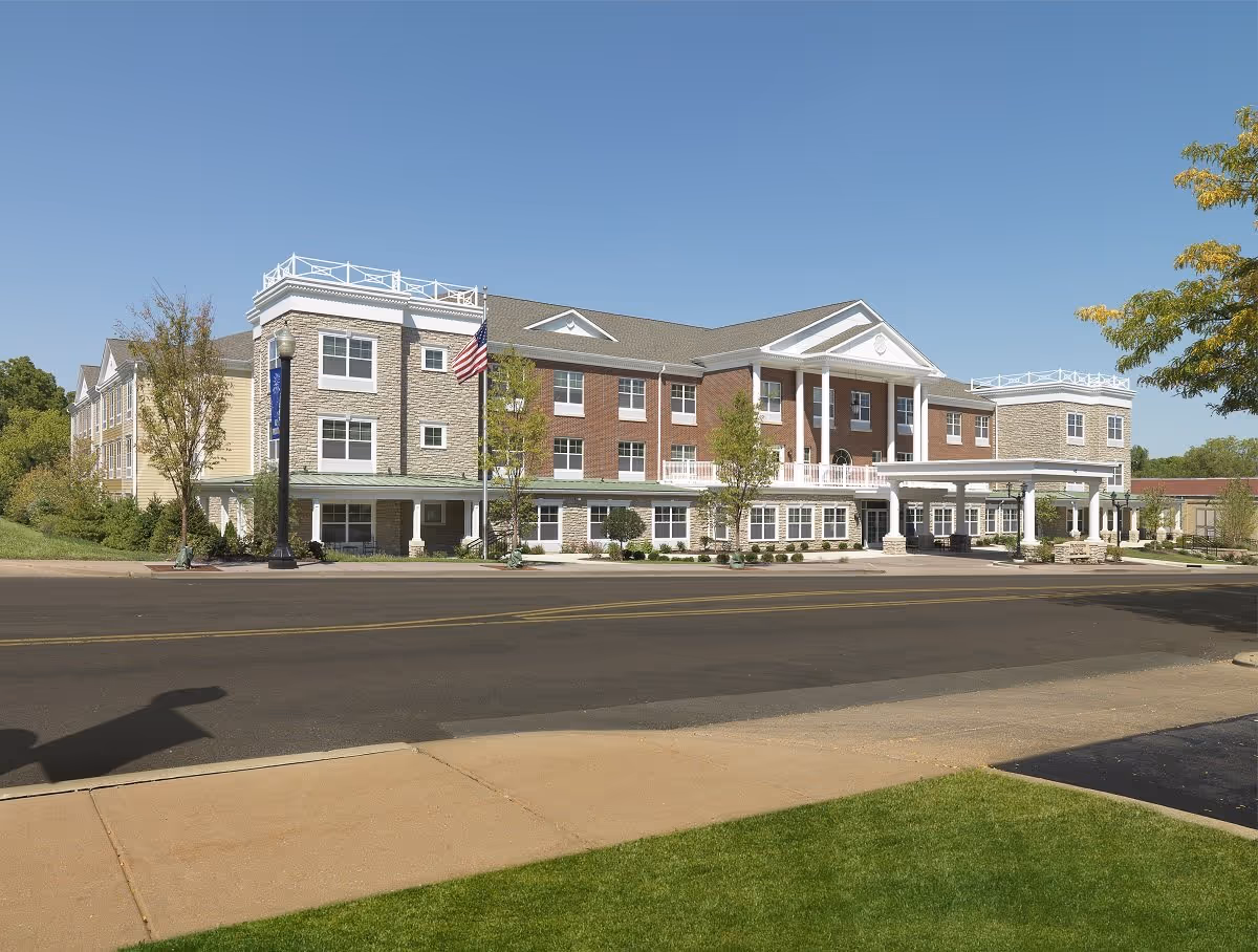 Exterior view of a large, multi-story senior living facility building with a combination of brick and stone facade, white columns, and a covered entrance. The building is surrounded by small trees and landscaping, with a clear blue sky overhead.