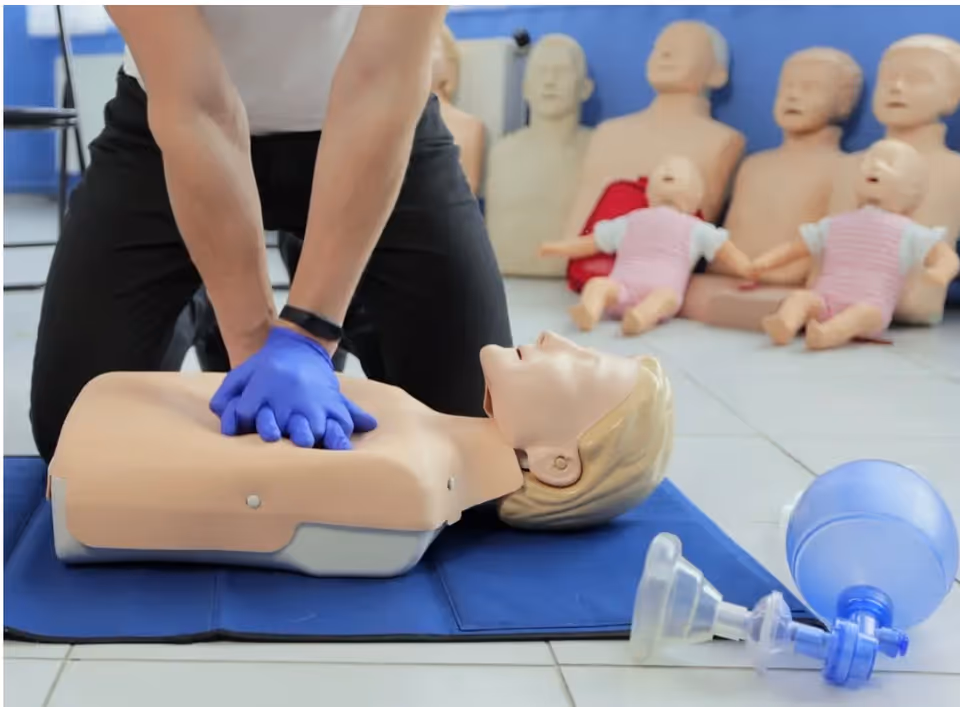 Gloved person performs chest compressions on a CPR dummy; several mannequins visible in background.