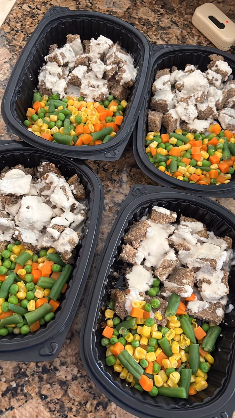 Four black meal trays on a granite counter, filled with diced vegetables and turkey with gravy.