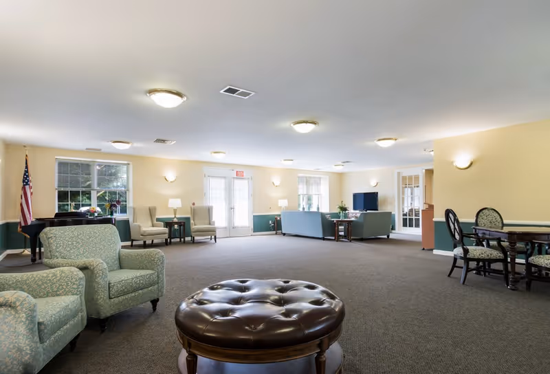 Spacious senior living lounge with teal upholstered chairs, round ottoman, piano, and multiple seating areas.