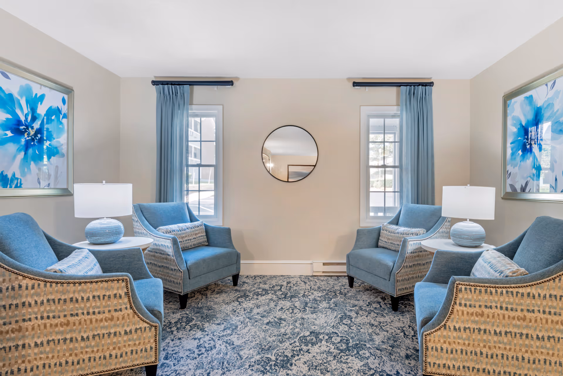 A cozy sitting area with four blue upholstered armchairs arranged in a square around two small white side tables, each holding a blue and white table lamp. The room features two windows with blue curtains, a round mirror centered on the wall between the windows, and two large framed blue floral artworks on the adjacent walls. The floor is covered with a blue and white patterned rug.