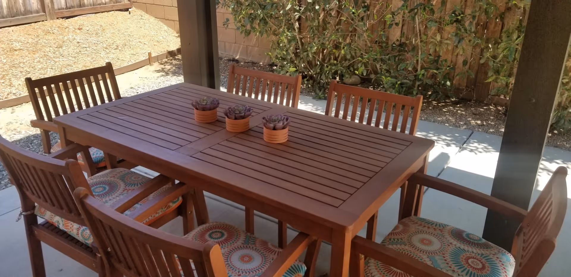 Outdoor patio area with a wooden table and six wooden chairs with colorful patterned cushions. Three small potted plants are placed in the center of the table. The patio is shaded by a roof and surrounded by a wooden fence and some greenery.