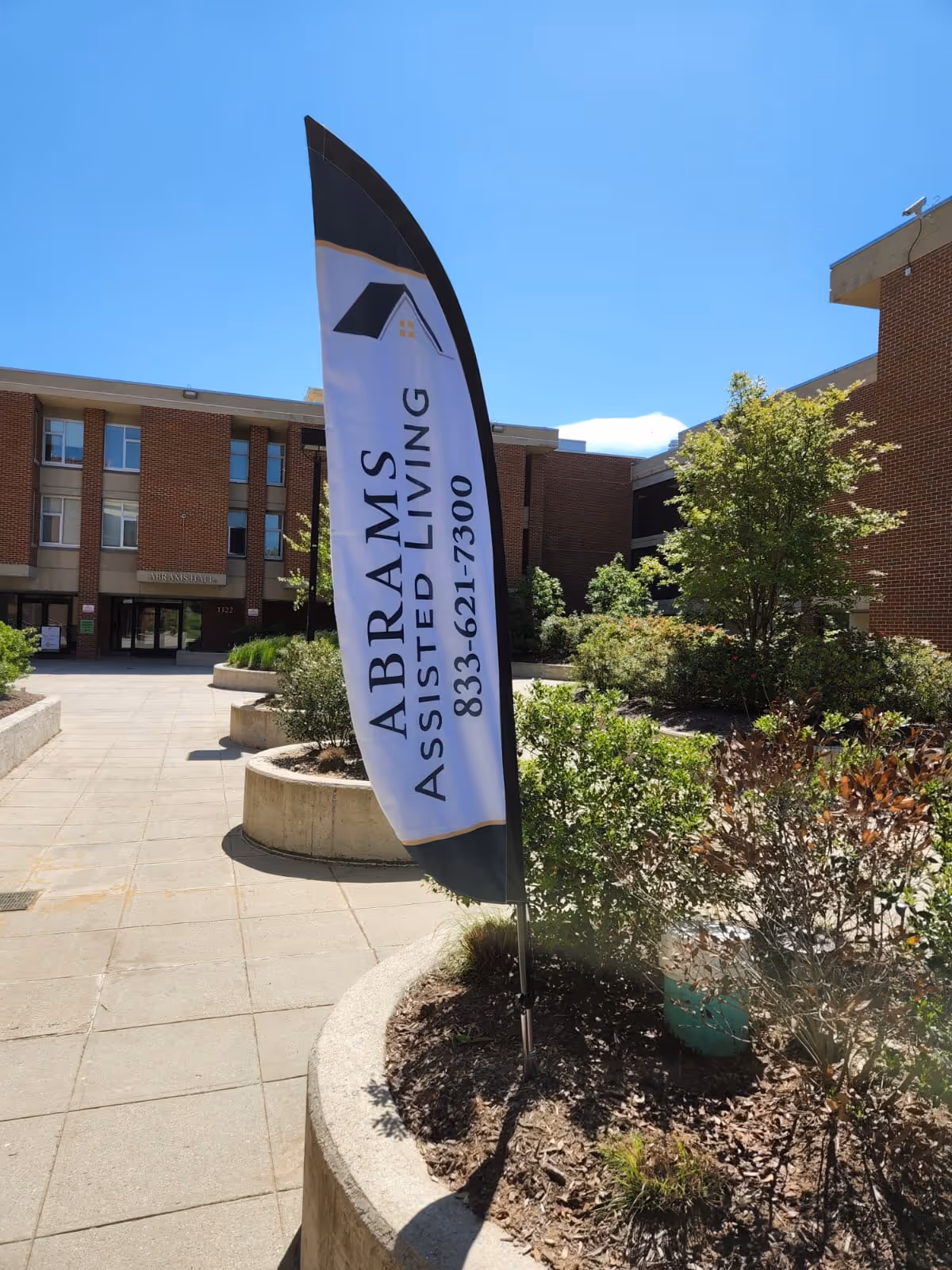 Brick senior apartment building exterior with a tall Abrams banner at a landscaped entrance.