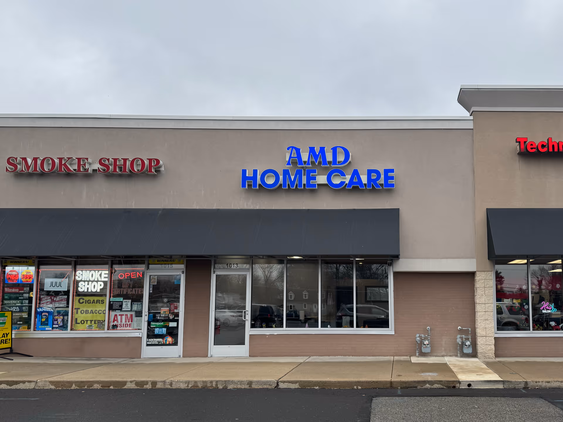 Exterior storefront with AMD Home Care signage in a strip mall beside a smoke shop