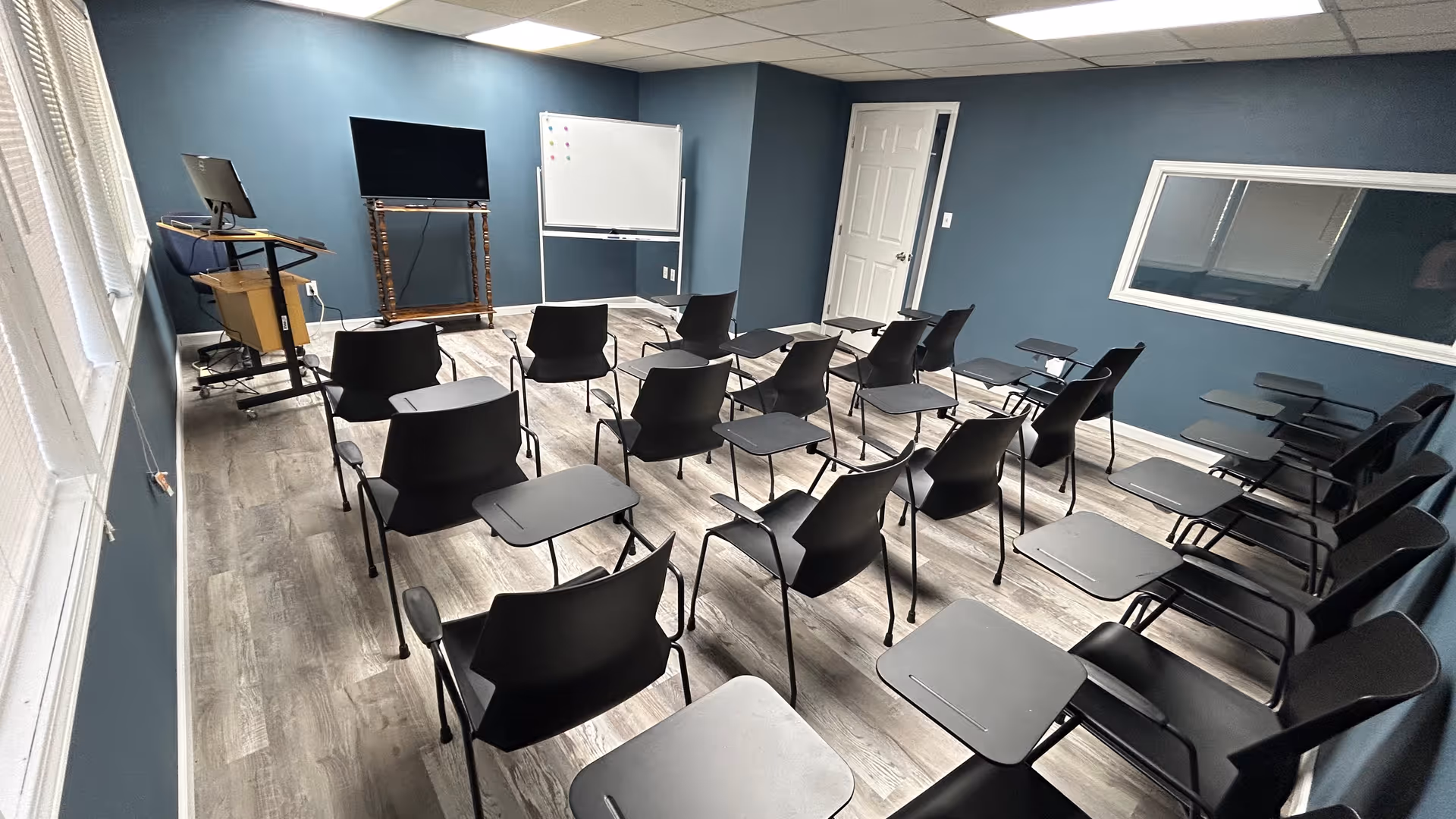 Empty training room with rows of black chairs and attached desks, whiteboard, and monitor.