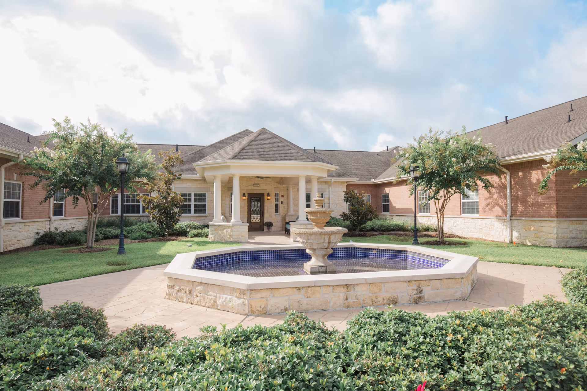 Front courtyard and entrance of a brick senior living building with a central fountain and surrounding landscaping.