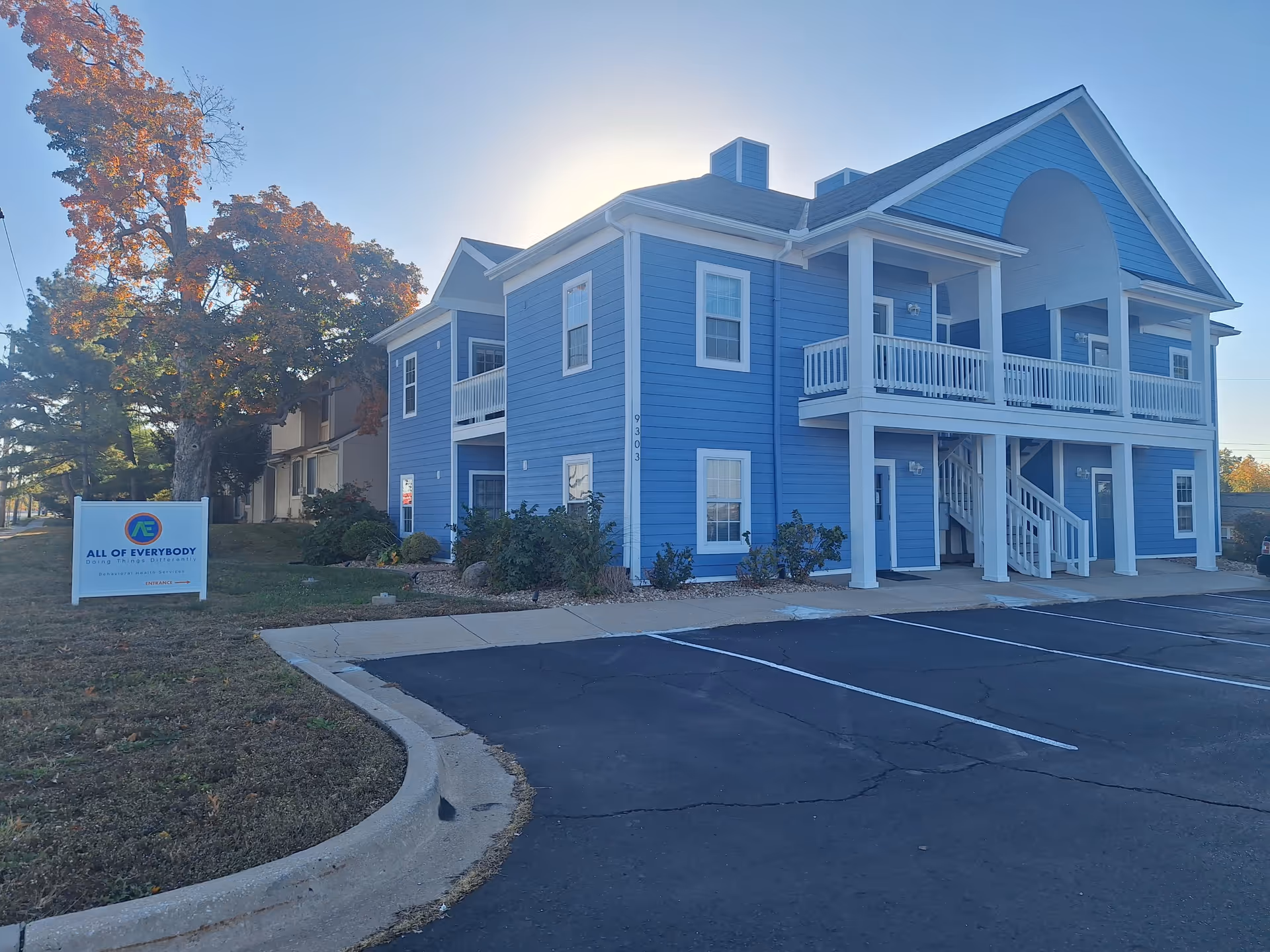 Blue two-story senior living building with white railings and a parking lot in front.
