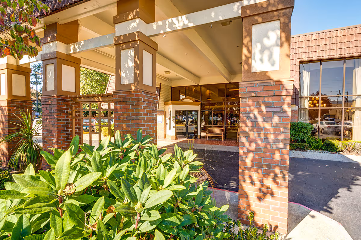 Covered main entrance of a senior living building with brick columns, glass doors, a bench, and landscaping in front.