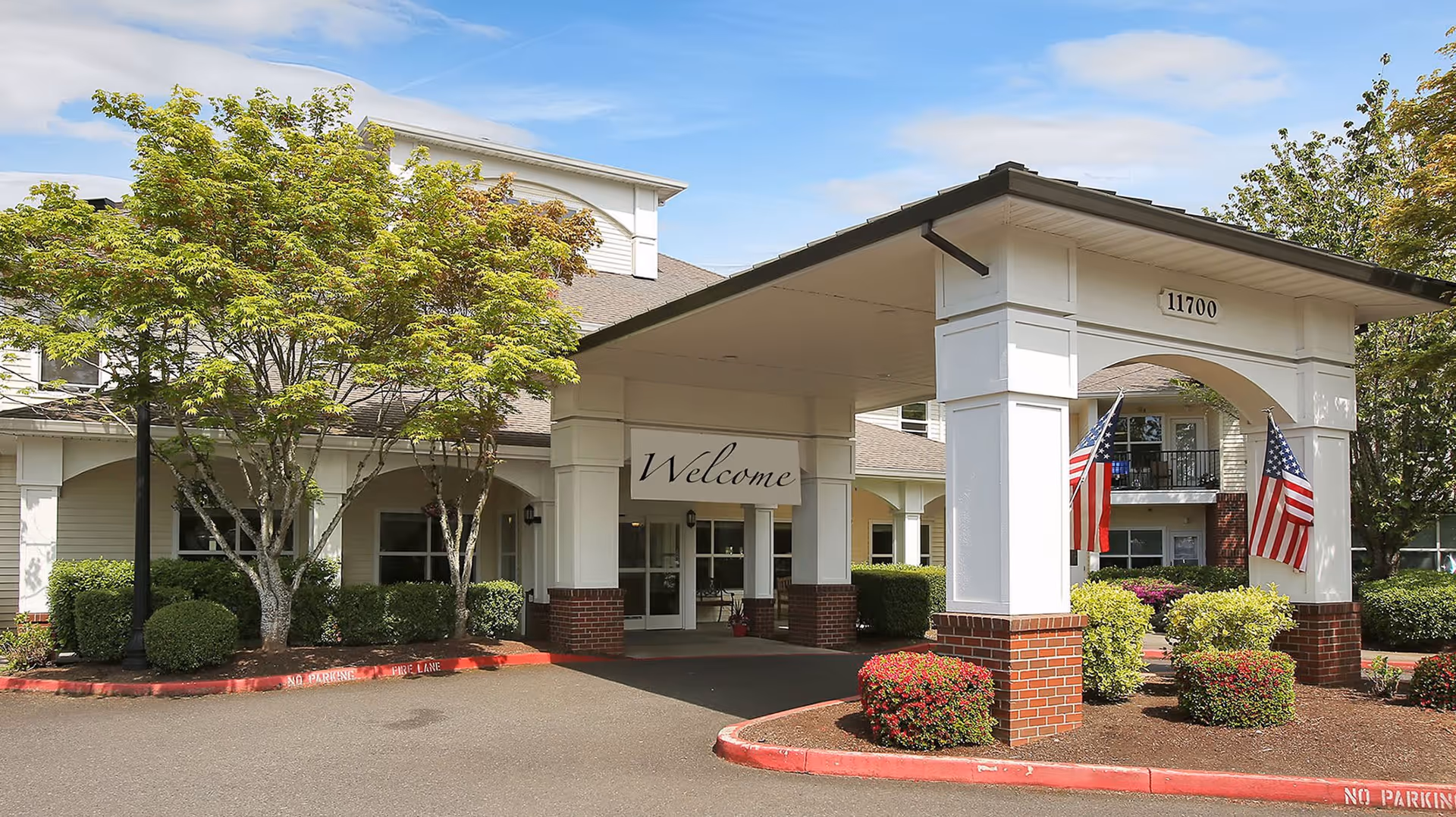 Entrance of a senior living facility with a covered drop-off area supported by white columns with brick bases. There are two American flags hanging on the columns, a 'Welcome' sign, and well-maintained landscaping including bushes and trees. The building number 11700 is displayed above the entrance.