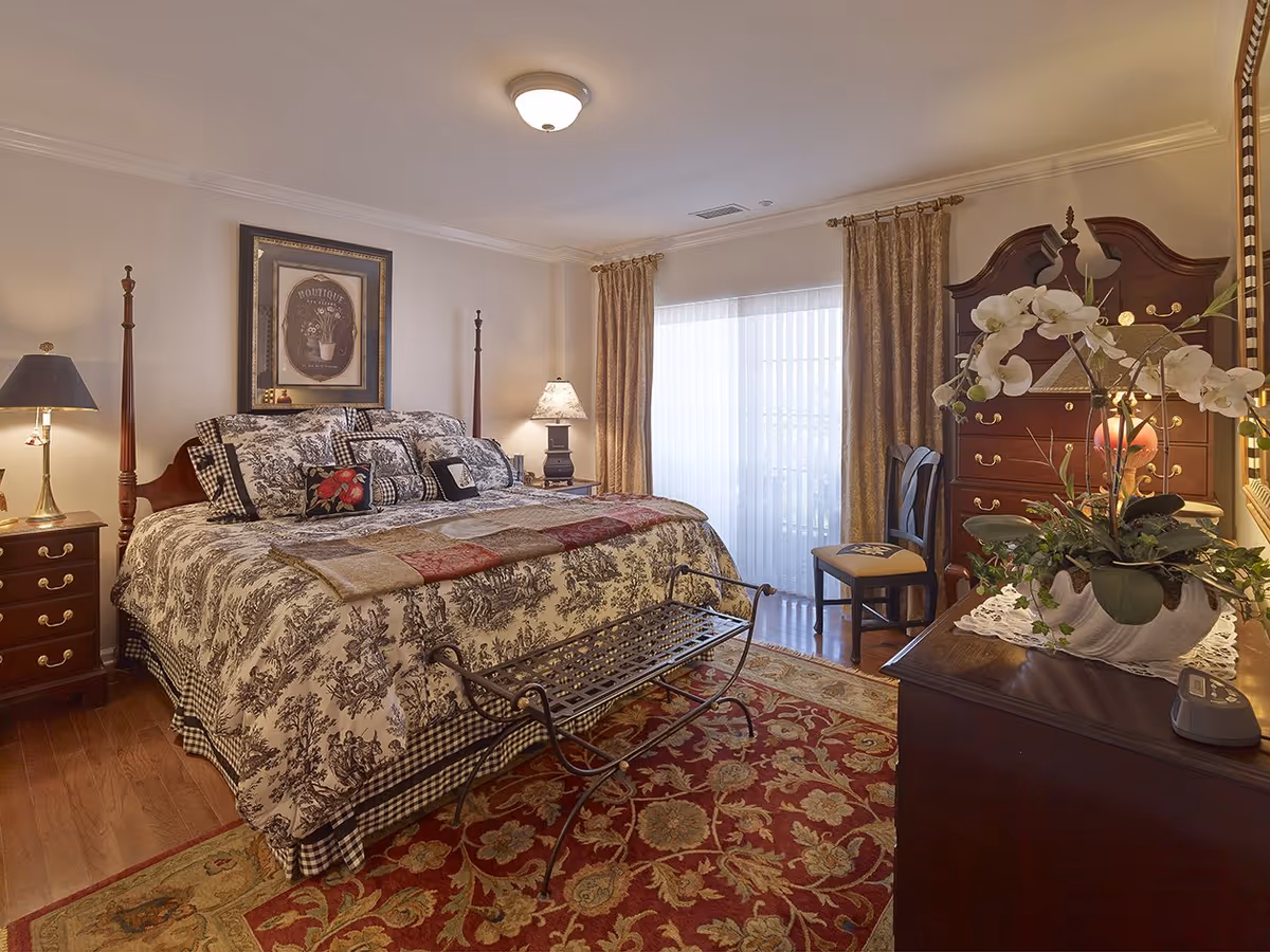 A well-decorated bedroom featuring a large bed with patterned bedding and multiple pillows. The room has wooden furniture including nightstands with lamps, a dresser with a mirror, a chair, and a decorative bench at the foot of the bed. There is a large window with sheer curtains and heavier drapes, and a floral arrangement on the dresser. The floor is wooden with a large patterned area rug.