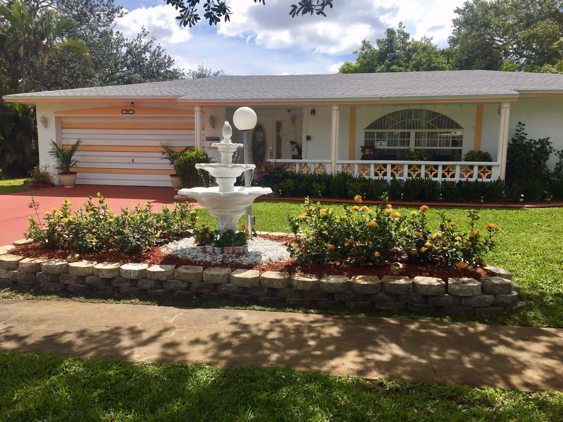 Front exterior view of a single-story assisted living facility with a gray roof, white walls, and peach-colored accents. The building features a garage door on the left, a front porch with decorative railing, and a large window with an arched top. In front of the building is a landscaped garden with a white tiered fountain, flowering bushes, and a red driveway.