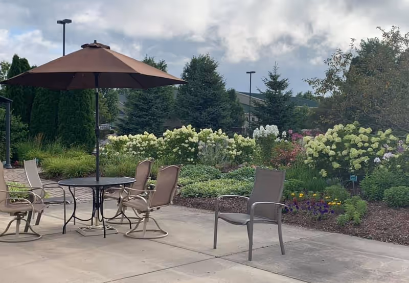 Outdoor patio with table, chairs, umbrella, and blooming garden beyond a paved courtyard.