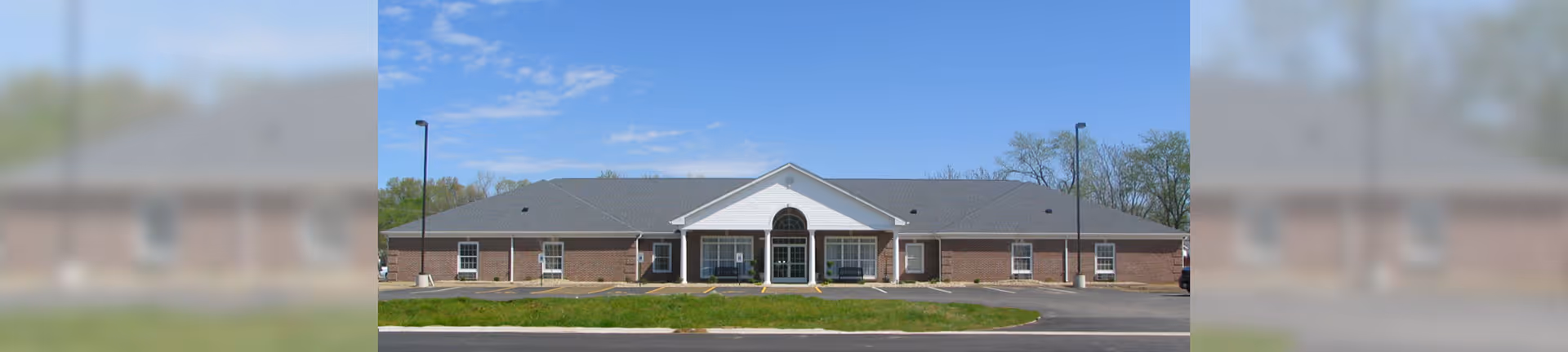 Single-story brick senior living building with a central covered entrance and pitched roof under a blue sky.