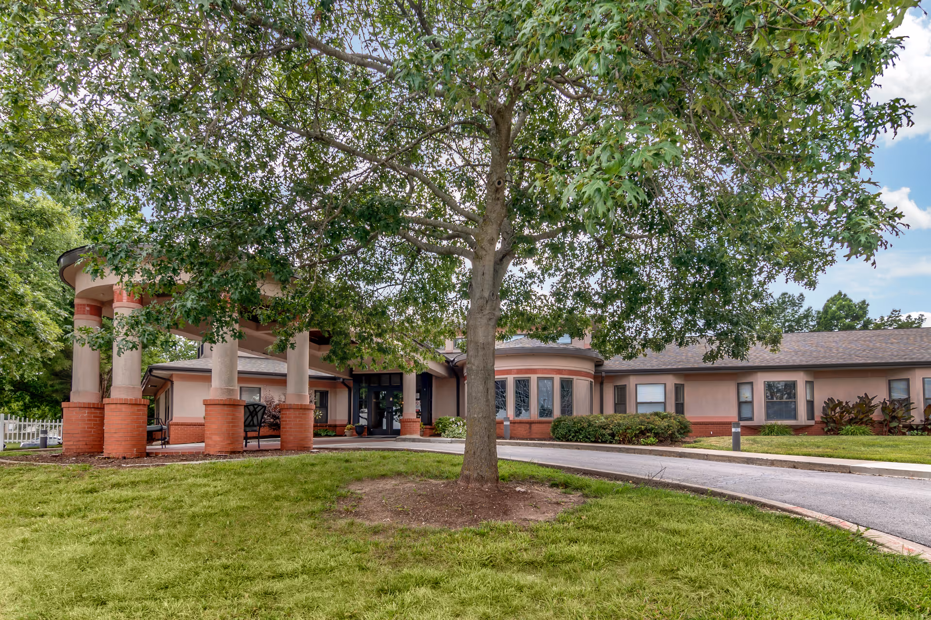 Front entrance of a single-story health care building with columns, a circular driveway, and a large tree in the foreground.