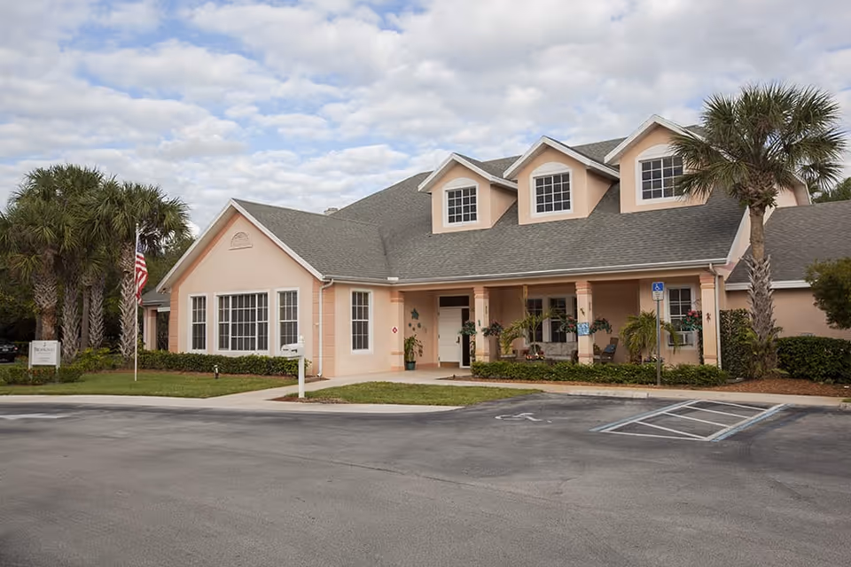 Front exterior of a peach-colored assisted living building with dormer windows, a covered entrance, palm trees, an American flag, and a parking lot with a handicap space.