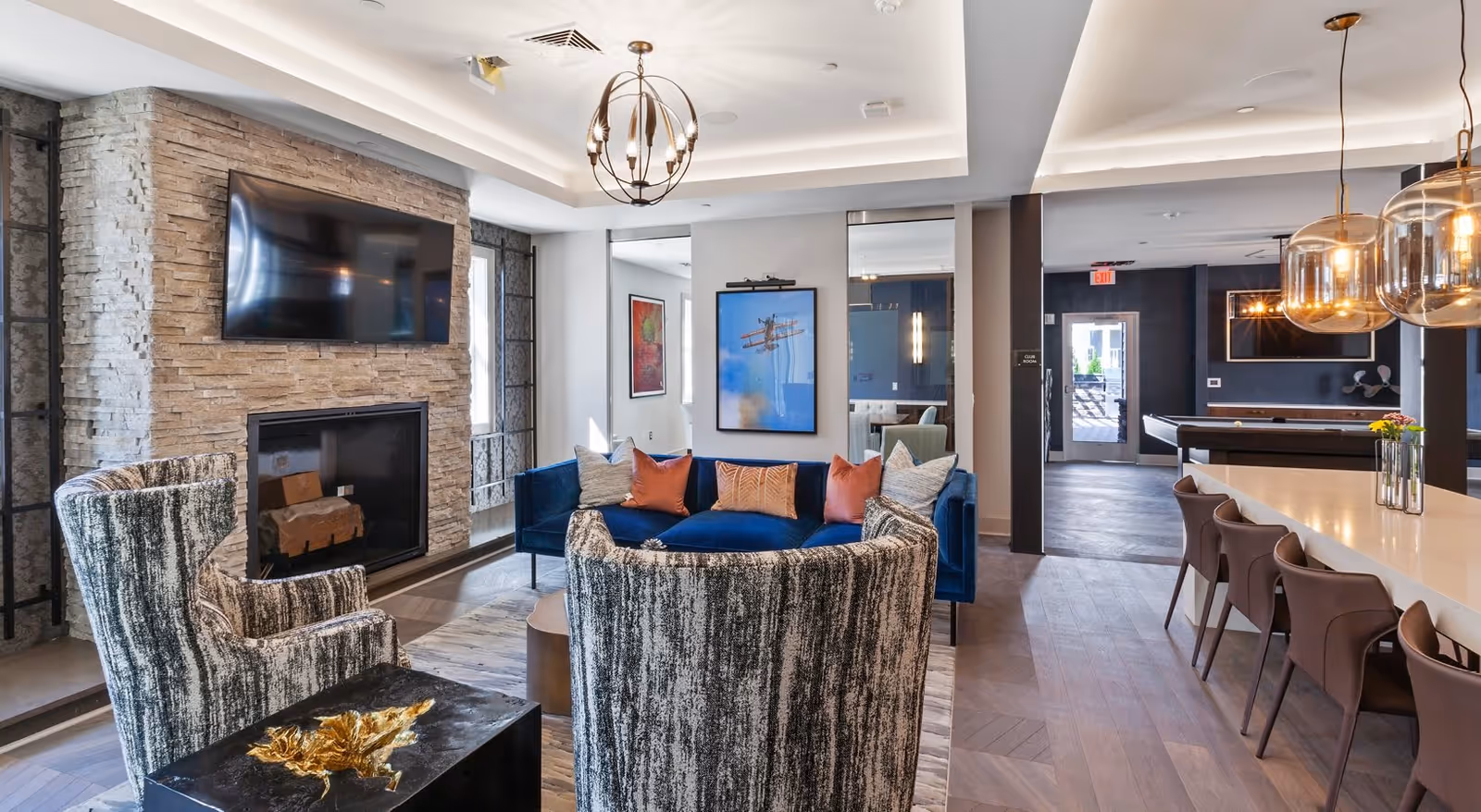 Bright modern common lounge with a stone fireplace and TV, blue sofa, patterned armchairs, and a long bar/dining counter.