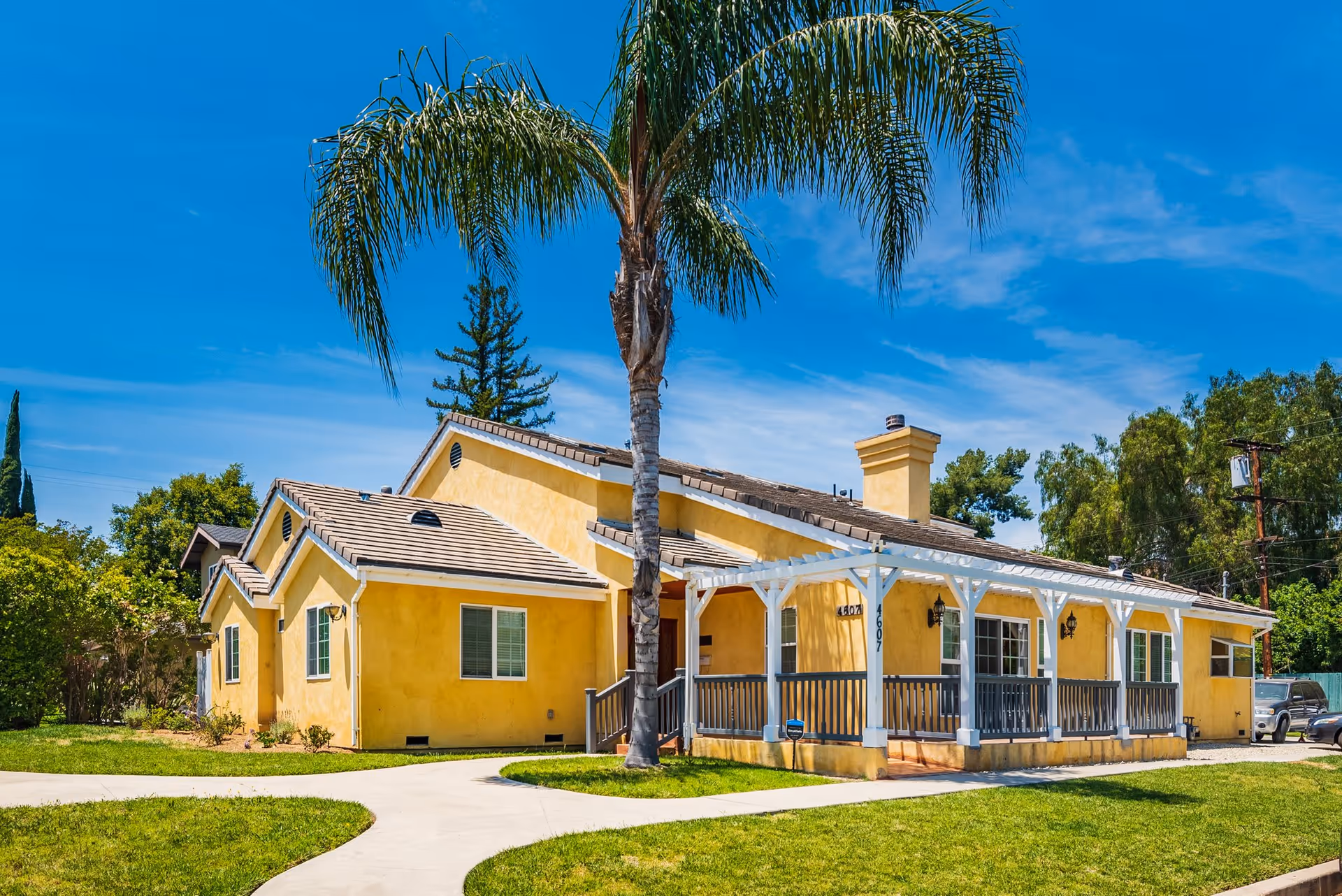 Exterior view of a single-story yellow building with a tiled roof and a white pergola over the front porch. There is a tall palm tree in front of the building, a green lawn, and a clear blue sky.