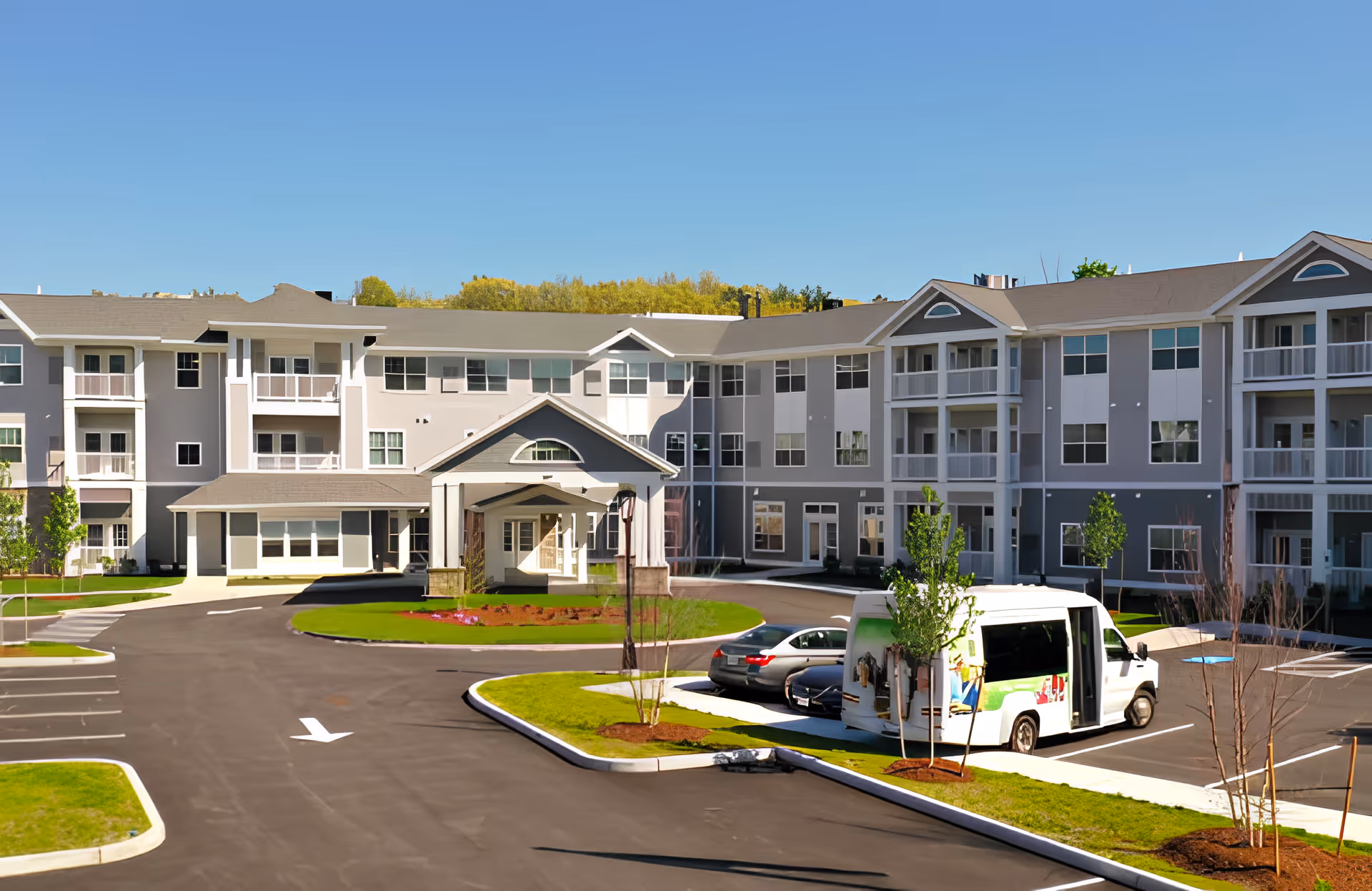 Exterior front view of a three-story retirement community building with a covered entrance, parking lot, and a shuttle van.