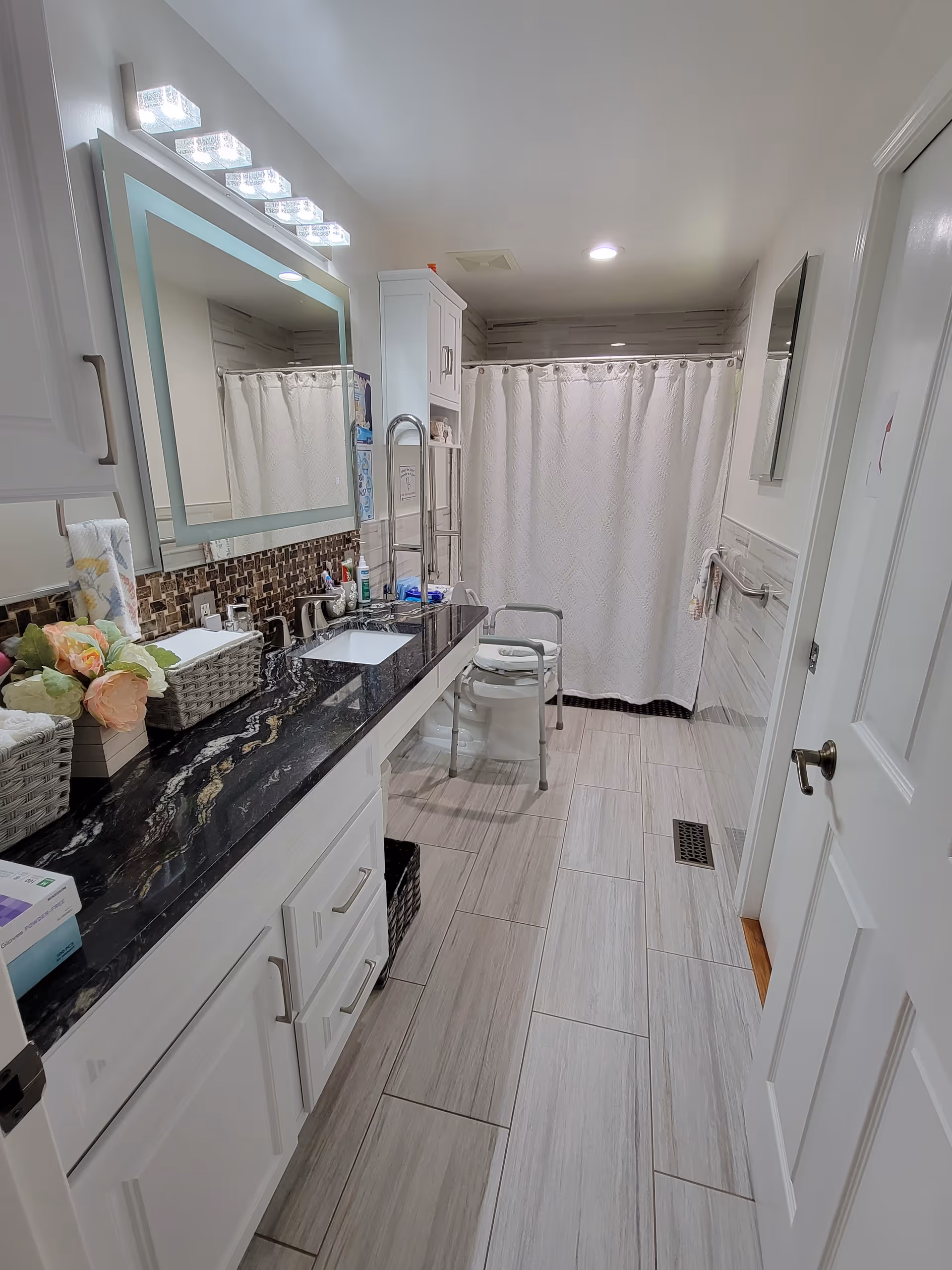 Accessible residential bathroom with a long black countertop, illuminated mirror, sink, grab bars, and a shower with curtain and shower chair.