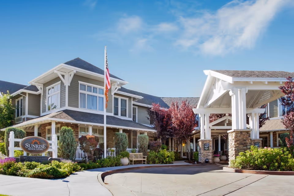 Exterior view of Sunrise of Sandy senior living facility showing a large two-story building with a covered entrance, landscaped greenery, an American flag, and a sign that reads 'Sunrise Senior Living'.