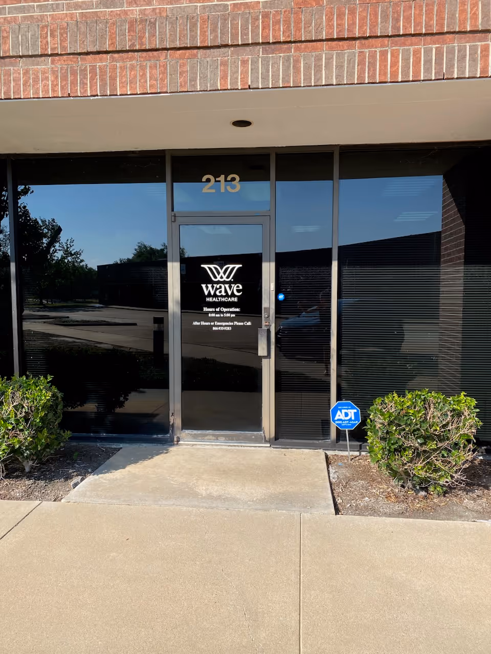 Glass entrance door with Wave Healthcare logo and 213 above, flanked by shrubs.