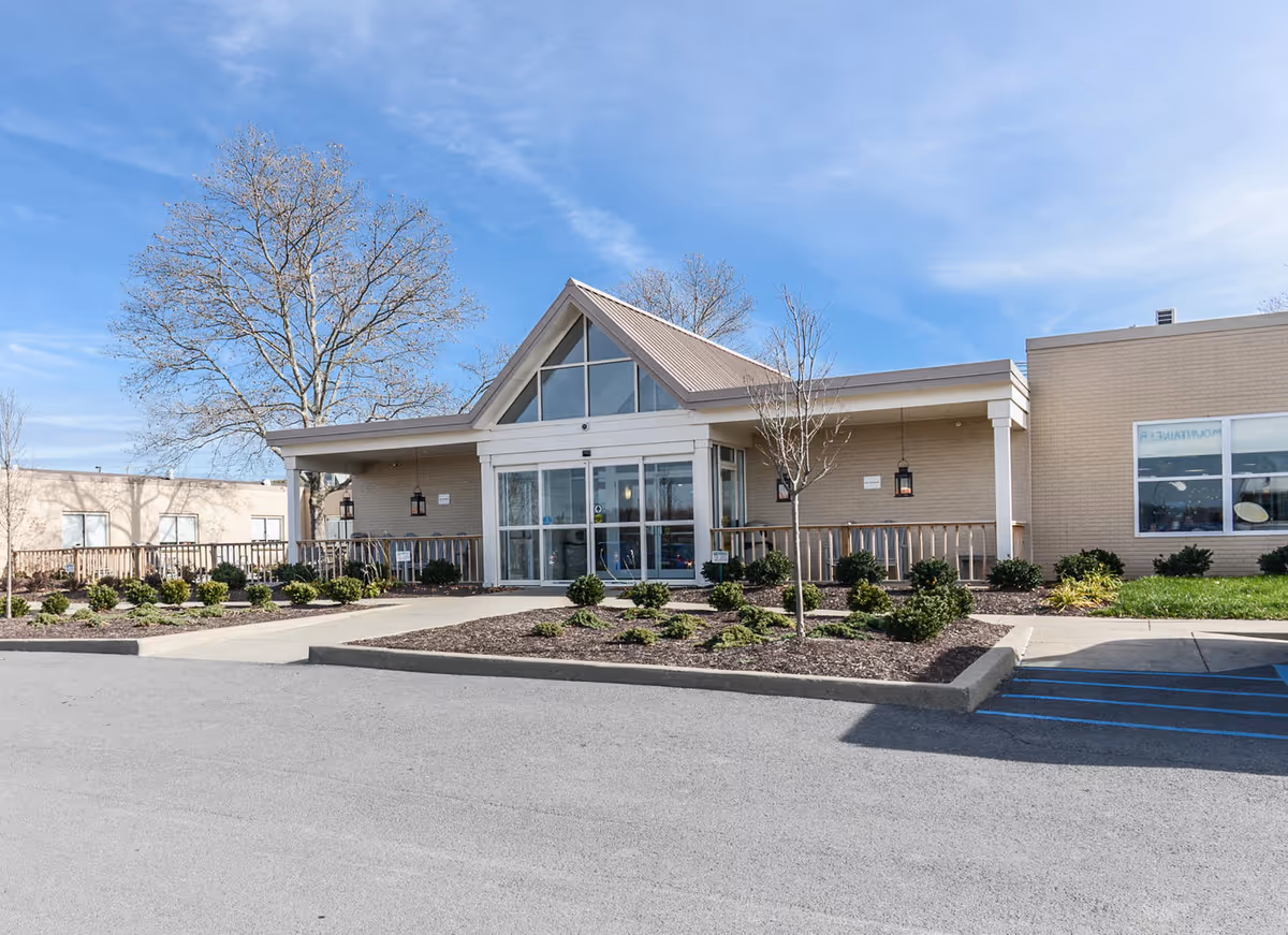 Front entrance of a single-story healthcare building with a triangular glass entry, landscaped beds, and a parking area.